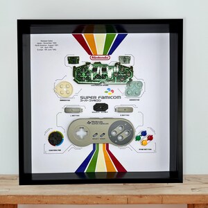 May include: A black shadow box frame containing a disassembled Super Famicom controller. The display features a green circuit board, various grey buttons, and the controller shell, all arranged on a white background with rainbow stripes and text.