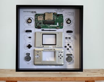 May include: A framed display featuring a deconstructed Nintendo DS. The components include the motherboard, speakers, buttons, and the console's front and back halves. The background is white with labels. The frame is black and sits on a wooden surface.
