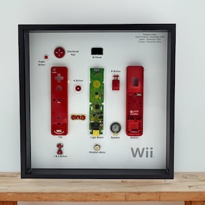 May include: A framed display featuring disassembled components of a Wii controller. The red controller parts, including the A and B buttons, are arranged with the logic board and speaker. The word "Wii" is displayed at the bottom.