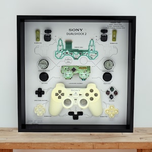 以下が含まれることがあります： PlayStation 2 DualShock 2 コントローラーの分解されたコンポーネントを特徴とする額入りのディスプレイ。コンポーネントは、ラベルと図が描かれた白い背景に配置されています。コントローラー自体は薄い黄色で、フレームは黒です。測定値は適用されません。