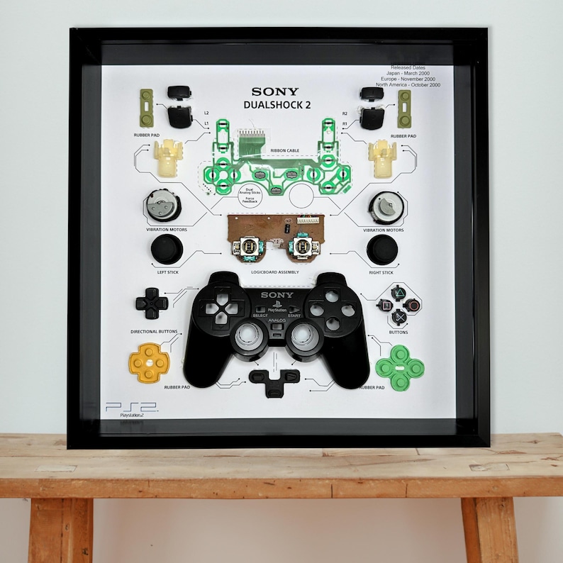 Pu&ograve; includere: Opera d'arte incorniciata con un controller Sony DualShock 2 smontato. Il controller nero &egrave; circondato dai suoi componenti, tra cui pulsanti, cuscinetti in gomma e circuiti stampati. La cornice &egrave; nera e lo sfondo &egrave; bianco.