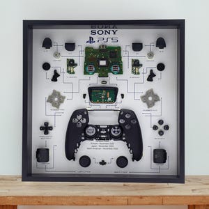 Könnte beinhalten: Gerahmte Darstellung eines zerlegten PlayStation 5-Controllers. Der schwarze Rahmen umgibt ein Diagramm der Komponenten des Controllers, einschließlich Tasten, Leiterplatten und des Controllers selbst. Der Text "SONY PS5" befindet sich oben.