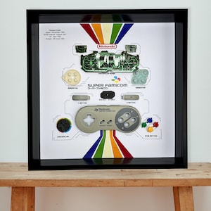 May include: Framed artwork showcasing a disassembled Super Famicom controller. The design includes a controller board, buttons, and the Super Famicom logo. The artwork is set against a white background with a rainbow stripe design and the Nintendo logo.