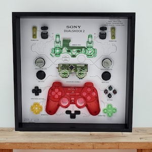 Könnte beinhalten: Gerahmtes Kunstwerk mit den zerlegten Komponenten eines Sony DualShock 2 Controllers. Das Design umfasst einen roten Controller, eine grüne Leiterplatte und verschiedene farbige Tasten und Pads, alle mit Fachbegriffen beschriftet.