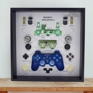 Könnte beinhalten: Gerahmtes Kunstwerk mit einem zerlegten Sony Dualshock 2 Controller. Der blaue Controller steht im Mittelpunkt, umgeben von beschrifteten Teilen wie Knöpfen, Gummipads und Leiterplatten. Der Rahmen ist schwarz, und der Hintergrund ist weiß.