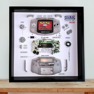 May include: A framed display of a disassembled Game Boy Advance. The components are laid out on a white background, with the screen showing "Pokémon Ruby Version." The black frame surrounds the components, with the text "GAME BOY ADVANCE".