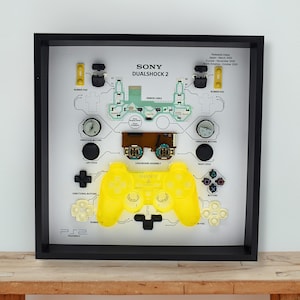 Puede incluir: Un marco que muestra un mando Sony Dualshock 2 desmontado. El mando amarillo es el punto central, con sus componentes dispuestos alrededor. El marco incluye el texto "SONY DUALSHOCK 2" e información técnica.