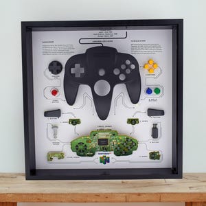 May include: A framed black shadow box art piece featuring a black Nintendo 64 controller. The artwork includes diagrams of controller components, a green circuit board, and the Nintendo 64 logo. The frame is displayed on a wooden surface.