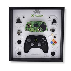 May include: Black framed display of an original Xbox controller and its components. The controller is black with the Xbox logo. The display includes the circuit board, buttons, and triggers, with text labels. The text reads "The First Generation Xbox Controller".