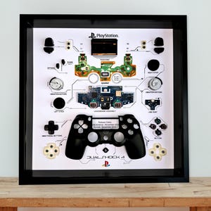 May include: A framed artwork displaying the disassembled components of a PlayStation DualShock 4 controller. The black controller is centered, with labeled parts like buttons, vibration motors, and circuit boards arranged around it. The frame is black, and the background is white.