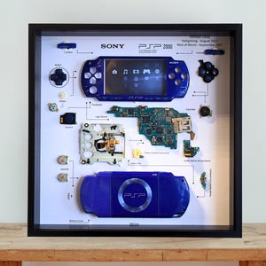 May include: A framed display of a disassembled blue Sony PSP 2000 handheld gaming console. The components are arranged to show the internal workings. Text includes "SONY PSP 2000" and release dates. The display is in a black frame.