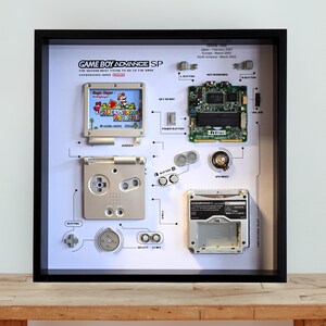 May include: A framed display of a disassembled Game Boy Advance SP, showing its components. The display includes the screen, motherboard, buttons, and a game cartridge with a Super Mario Advance game. The text "GAME BOY ADVANCE SP" is visible.