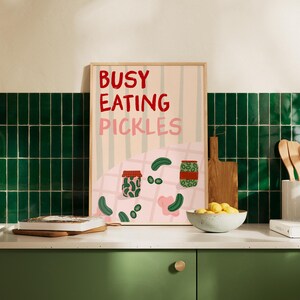 May include: A framed art print with the text "BUSY EATING PICKLES" in red and pink. The print features illustrations of pickles and jars on a pink and white striped background. The print is displayed on a green countertop.