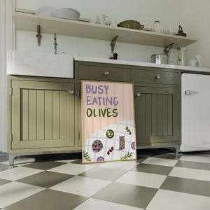 May include: A poster with the text "BUSY EATING OLIVES" in purple and green. The poster shows illustrations of olives and a checkered tablecloth. The poster is leaning against a green cabinet in a kitchen setting.