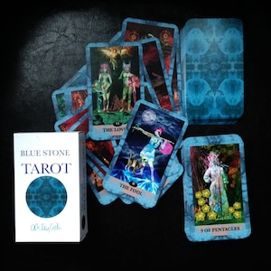Può includere: Un mazzo di tarocchi Blue Stone con una scatola bianca con la scritta "BLUE STONE TAROT" e un design circolare blu. Sono visibili diverse carte dei tarocchi, tra cui "Gli Amanti", "Il Matto" e "9 di Denari."