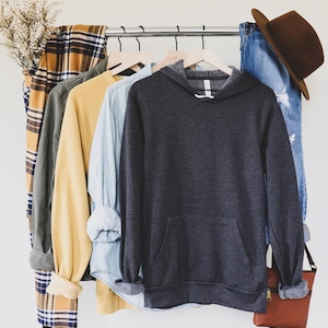 May include: A dark grey hooded sweatshirt with a front pocket hanging on a rack with other clothing items, including a plaid shirt, a green shirt, a yellow shirt, and a light blue shirt.