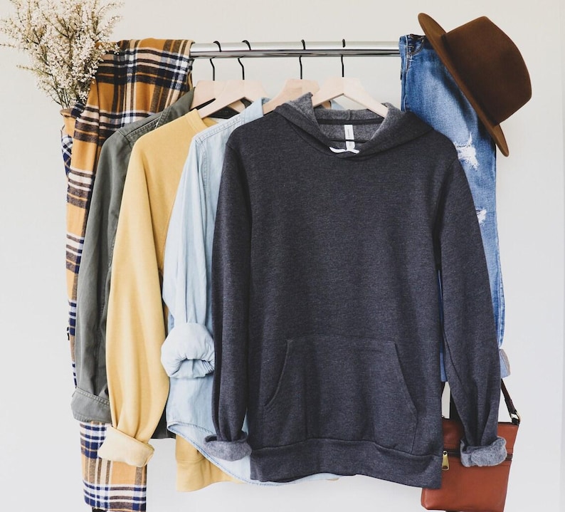 May include: A dark grey hooded sweatshirt with a front pocket hanging on a rack with other clothing items, including a plaid shirt, a green shirt, a yellow shirt, and a light blue shirt.