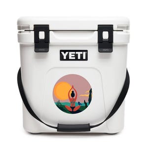 May include: White YETI cooler with a black strap and a round sticker featuring a woman doing yoga in front of a sunset. The sticker has a pink, orange, and blue color scheme.
