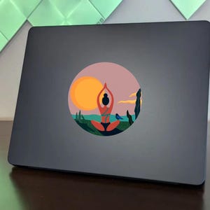 May include: A round sticker with a colorful illustration of a person in a yoga pose, a sunset, and a bird. The sticker is on a gray laptop.