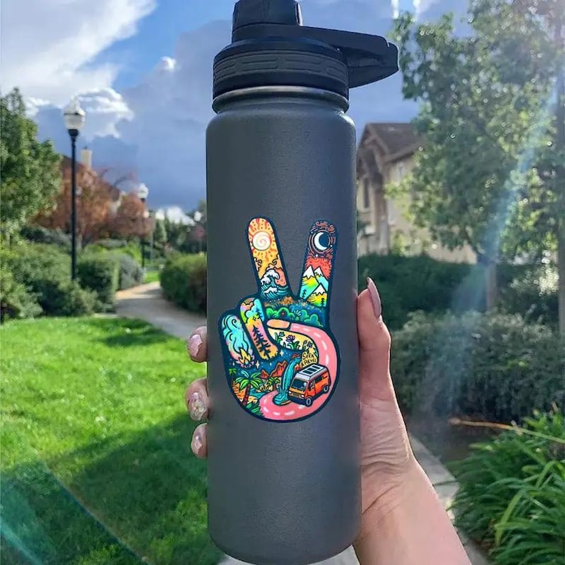 May include: A gray reusable water bottle with a peace sign sticker. The sticker features a colorful design with a van, mountains, a sun, and a moon.