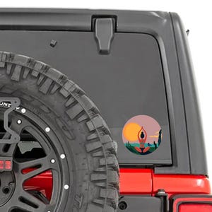 May include: A red Jeep with a black spare tire and a sticker on the back window. The sticker is a circle with a sunset scene and a person in a yoga pose.