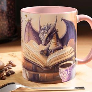 Puede incluir: Una taza de cer&aacute;mica rosa con un drag&oacute;n leyendo un libro. El drag&oacute;n es morado y tiene alas. El libro est&aacute; abierto y tiene una pluma en la parte superior. La taza est&aacute; rodeada de una pila de libros.