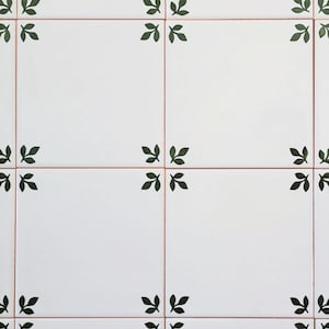 Può includere: Piastrelle in ceramica bianca con un disegno di foglie verdi in ogni angolo. Le piastrelle sono disposte a griglia, con sottili linee marroni che separano ogni piastrella. Le foglie sono di colore verde scuro.