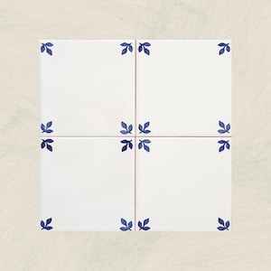 Può includere: Quattro piastrelle di ceramica bianche disposte a quadrato, ognuna ornata da un motivo a foglie blu negli angoli. Le piastrelle sono poste su uno sfondo neutro, mostrando un'estetica pulita e minimalista, adatta all'arredamento della casa.
