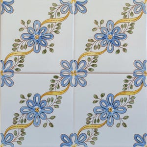 May include: Four white ceramic tiles arranged to create a repeating pattern. Each tile features a blue flower with yellow ribbon-like accents and green leaves. The floral design is centered on each tile.