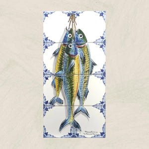 May include: Three blue and yellow fish with black eyes are hanging from a string on a white tile with a blue and white floral pattern. The tile is part of a larger set of tiles.