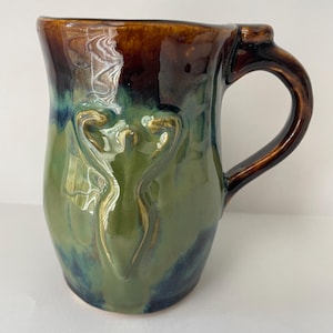 May include: A handmade ceramic mug with a glossy finish. The mug features a dark brown rim, transitioning to green and blue hues on the body. Two stylized, gold-colored figures are embossed on the front. The handle is a dark brown color.