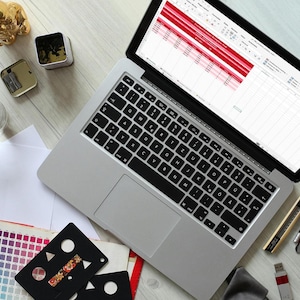May include: A silver laptop displaying a spreadsheet with red highlighted rows, surrounded by a pencil case labeled TOOL KIT, a color swatch book, a black stencil, a gold figurine, a glass of water, and various writing instruments on a desk.