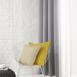 May include: A white wire chair with a yellow and beige cushion. The chair is in front of a white brick wall and grey curtains.