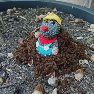 Può includere: Una talpa all'uncinetto con un casco giallo e una salopette blu spunta da un mucchio di lana marrone.