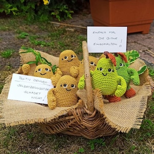 Könnte beinhalten: Ein Weidenkorb gefüllt mit handgefertigten Häkelfiguren, darunter gelbe kartoffelartige Charaktere und grüne birnenförmige Figuren. Der Korb steht auf Jute und Gras, mit Schildern auf Deutsch. Die Figuren haben lächelnde Gesichter.