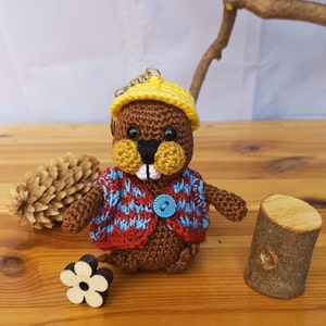 May include: A handmade crochet beaver keychain. The beaver is brown with a yellow hat and a blue and red patterned vest. It has a black nose, yellow cheeks, and a blue button. The keychain is on a wooden surface.