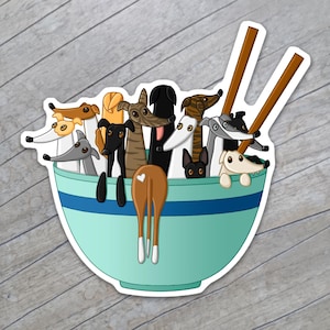 Op de afbeelding: Een sticker met een turquoise kom met een blauwe streep, gevuld met cartoon windhonden. Bruine eetstokjes zijn aan de zijkant geplaatst. De windhonden zijn in verschillende kleuren, waaronder bruin, zwart en wit. De achterkant van een windhond is zichtbaar.