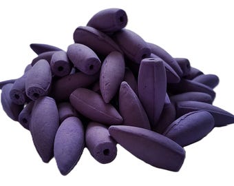 Peut inclure: Un tas de cônes d'encens violets, chacun présentant une surface mate et texturée avec un petit trou circulaire au sommet. Les cônes ont une forme de balle arrondie et allongée, empilés en un amas dense et irrégulier sur un fond blanc.