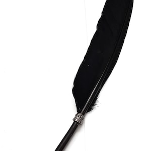 May include: A black feather quill pen featuring a long, dark plume attached to a slender black shaft. The pen is accented with two ornate, silver-toned metal bands with intricate embossed detailing, and it terminates in a pointed metal nib.