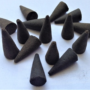 Pode incluir: Uma coleção de cones de incenso castanhos escuros, cada um com formato cónico e superfície texturizada. Os cones estão espalhados sobre uma superfície branca, criando um arranjo visualmente atraente. Estes cones de incenso são projetados para aromaterapia e relaxamento.