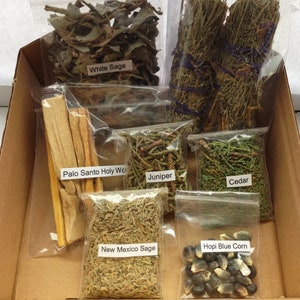 Puede incluir: Una caja de cartón llena de varias hierbas y productos botánicos secos. La caja contiene bolsas de plástico transparente etiquetadas como "White Sage", "Palo Santo Holy Wood", "Juniper", "Cedar", "New Mexico Sage" y "Hopi Blue Corn". Los manojos de salvia están atados con hilo morado.