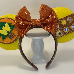 May include: Yellow Minnie Mouse ears with a brown headband. One ear features a brown and orange felt patch with a green tree and the letter W. The other ear features a brown felt patch with gold glitter and several small, colorful, felt patches.
