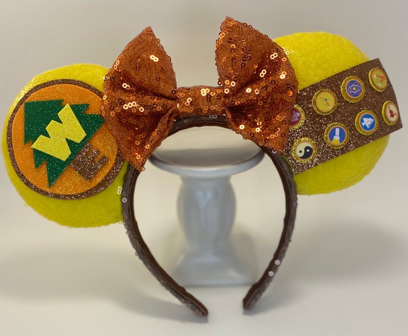May include: Yellow Minnie Mouse ears with a brown headband. One ear features a brown and orange felt patch with a green tree and the letter W. The other ear features a brown felt patch with gold glitter and several small, colorful, felt patches.