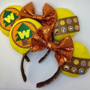 May include: A pair of yellow and brown Minnie Mouse ears with a brown bow and a yellow and brown design featuring a tree and the letter W. The ears are decorated with small, round, colorful buttons.