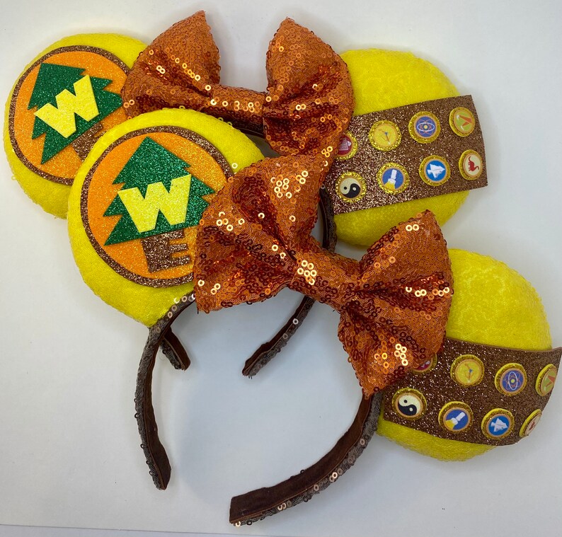 May include: A pair of yellow and brown Minnie Mouse ears with a brown bow and a yellow and brown design featuring a tree and the letter W. The ears are decorated with small, round, colorful buttons.