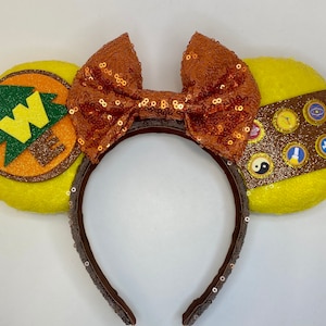 May include: Yellow mouse ears with a brown headband, a brown bow with orange sequins, and a brown patch with a green tree and the letters "WE" on one ear. The other ear has a brown patch with gold glitter and several small gold badges with different symbols.