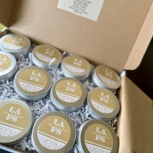May include: A box filled with small, round, silver tins. Each tin has a gold label with the letters "LA FS" and a decorative border. The box also contains a list of scents. The tins are nestled in white shredded paper.