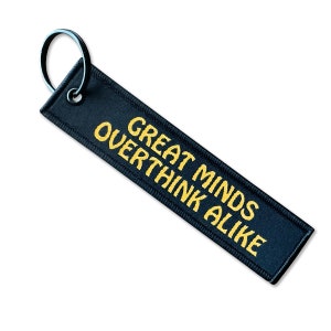 May include: Black fabric keychain with a gold embroidered saying "Great Minds Overthink Alike".