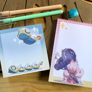 Könnte beinhalten: Zwei quadratische Notizblöcke mit Cartoon-Illustrationen. Einer ist blau mit einem fliegenden Wesen und einem segmentierten Wurm, der andere rosa mit einem Roboter und einem rosa Wesen. Stifte und Würfel sind ebenfalls zu sehen.