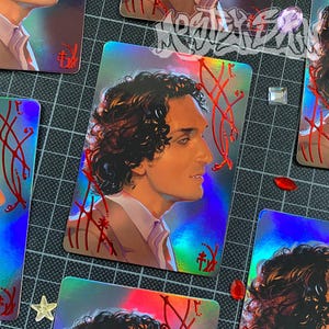 Könnte beinhalten: Eine rechteckige holografische Sammelkarte mit einer Profilillustration einer Person mit dunklem, lockigem Haar und einem hellen Hemd mit Kragen. Die Kartenoberfläche zeigt einen reflektierenden Regenbogen-Schimmer und rote Linienverzierungen.